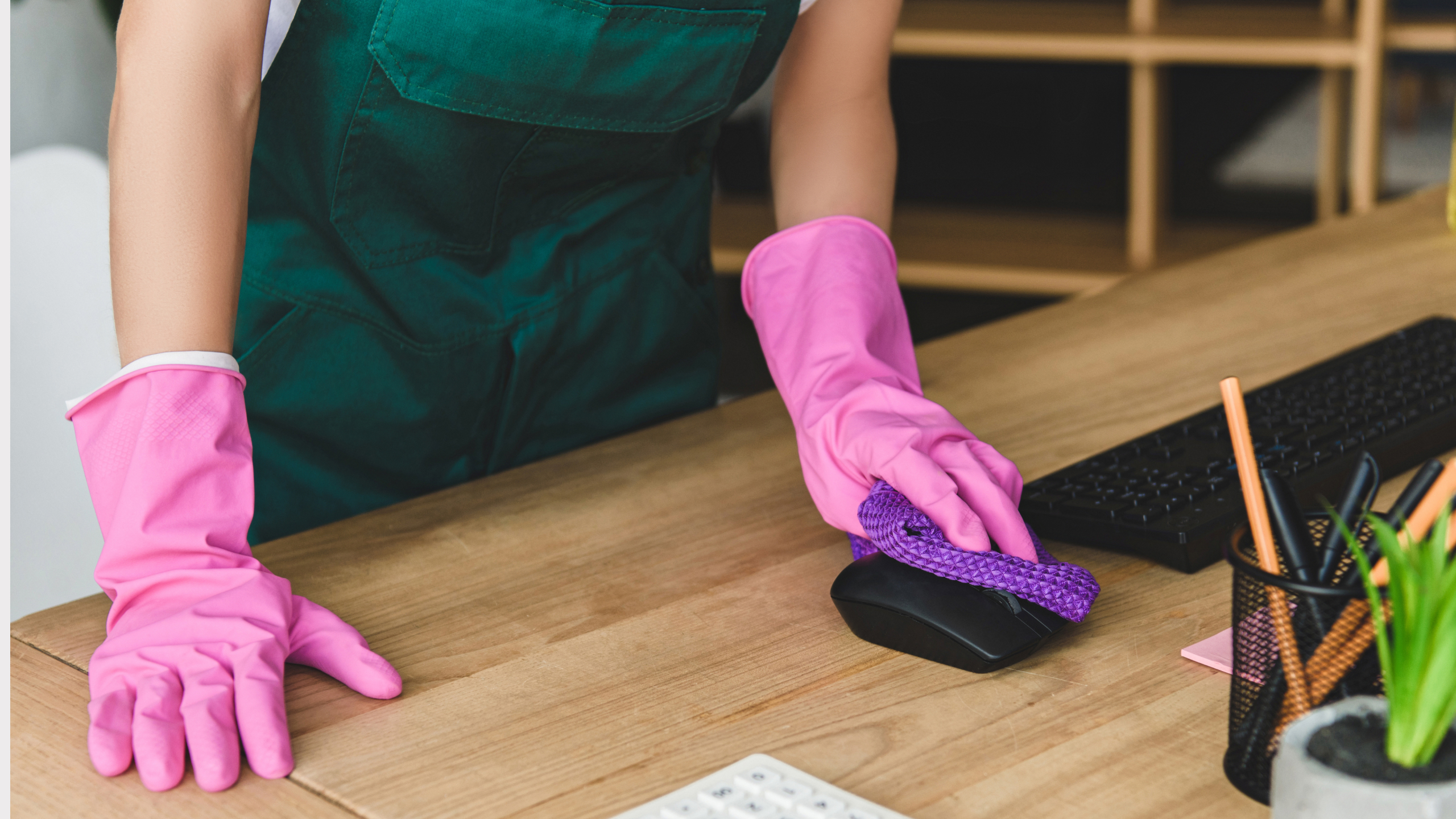 A person wearing pink rubber gloves is cleaning a wooden desk with a purple cloth, near a computer mouse and various office supplies.